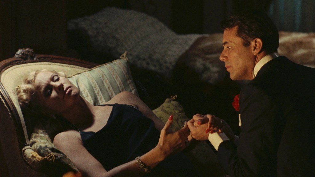 A man in a suit holds the hand of a reclining woman on a chaise longue,